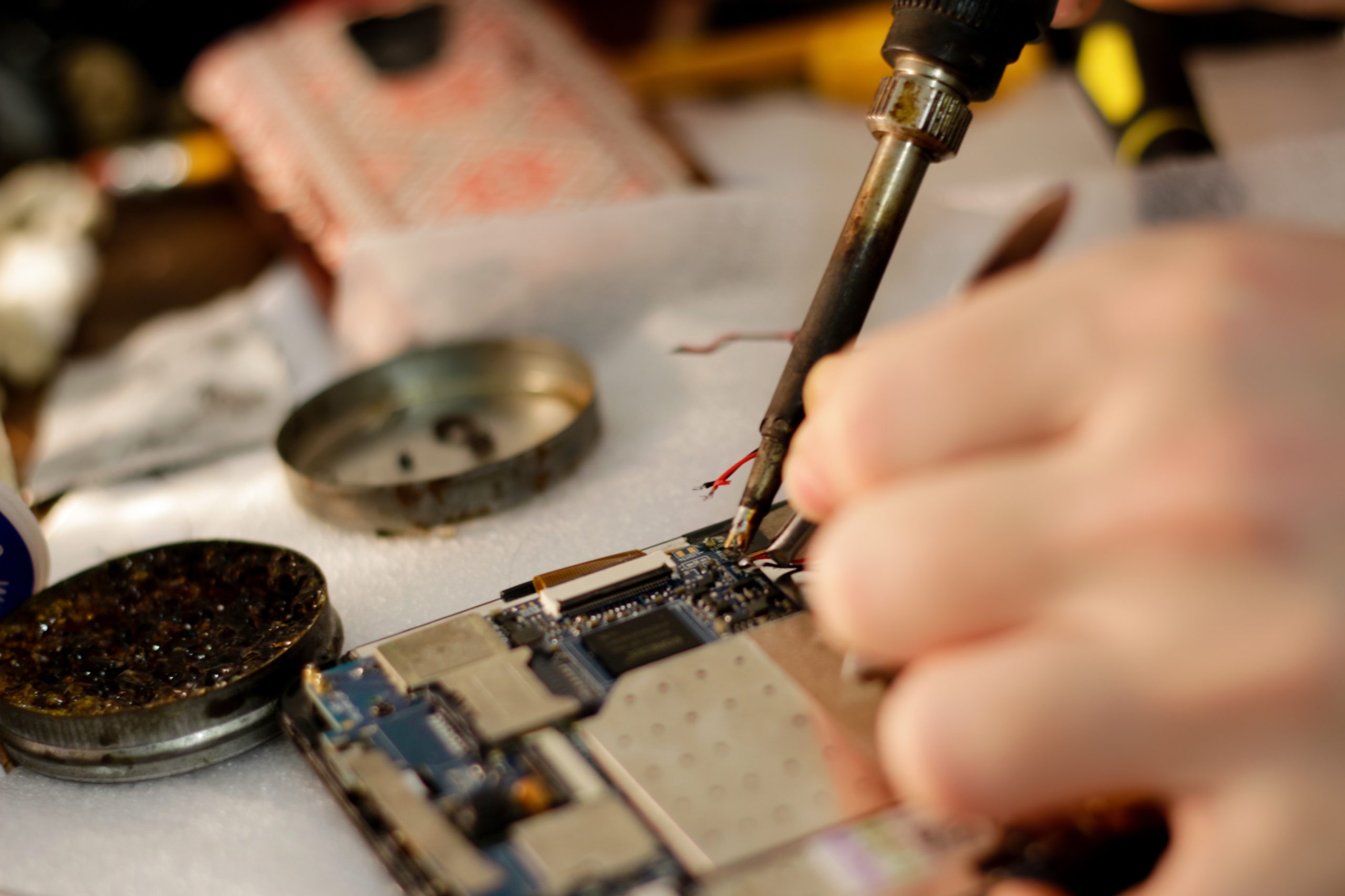 A close-up of a person soldering wires onto a smartphone circuit board with a soldering iron, illustrating the right to repair. Various tools and materials are visible in the background.