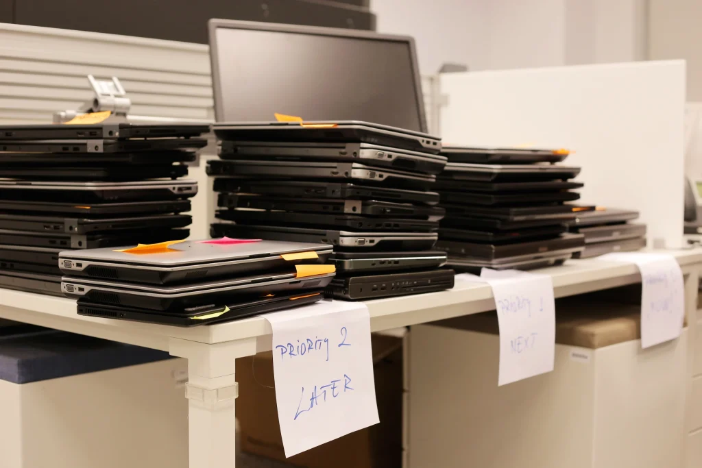 Several stacks of closed laptops with colored sticky notes are placed on a white table, resembling the organized system of orders in a restaurant. Handwritten labels under the stacks read “priority 2 Later,” “priority 1 Next,” and “Ready.”.