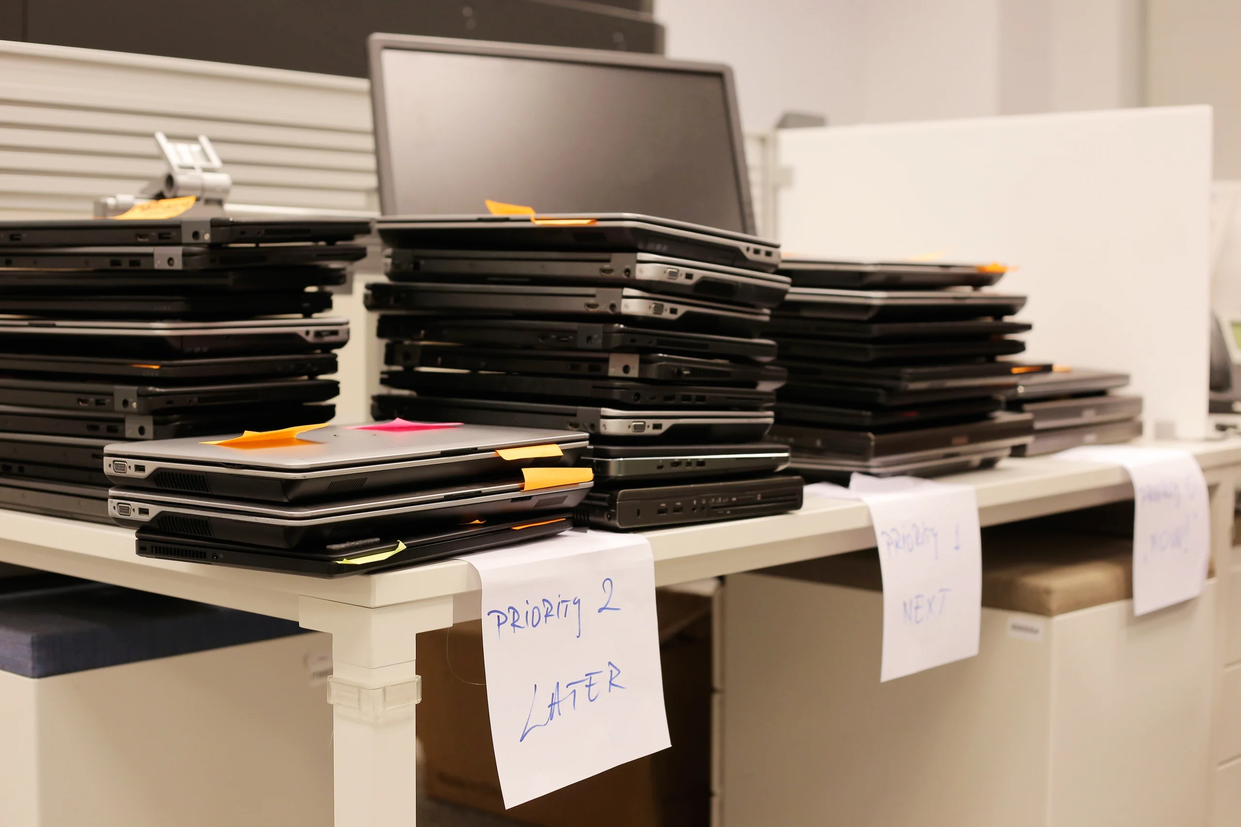 Several stacks of closed laptops with colored sticky notes are placed on a white table, resembling the organized system of orders in a restaurant. Handwritten labels under the stacks read “priority 2 Later,” “priority 1 Next,” and “Ready.”.