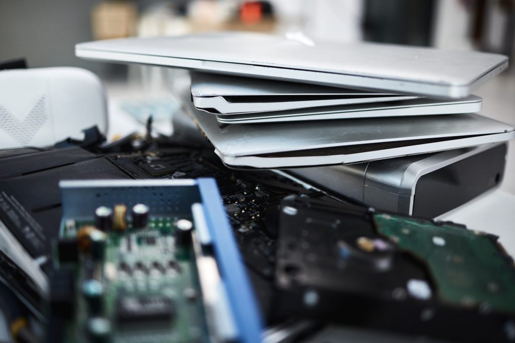 A pile of old electronic devices, including several stacked laptops and scattered computer parts like circuit boards and hard drives, highlights potential cybersecurity risks if not properly disposed of.