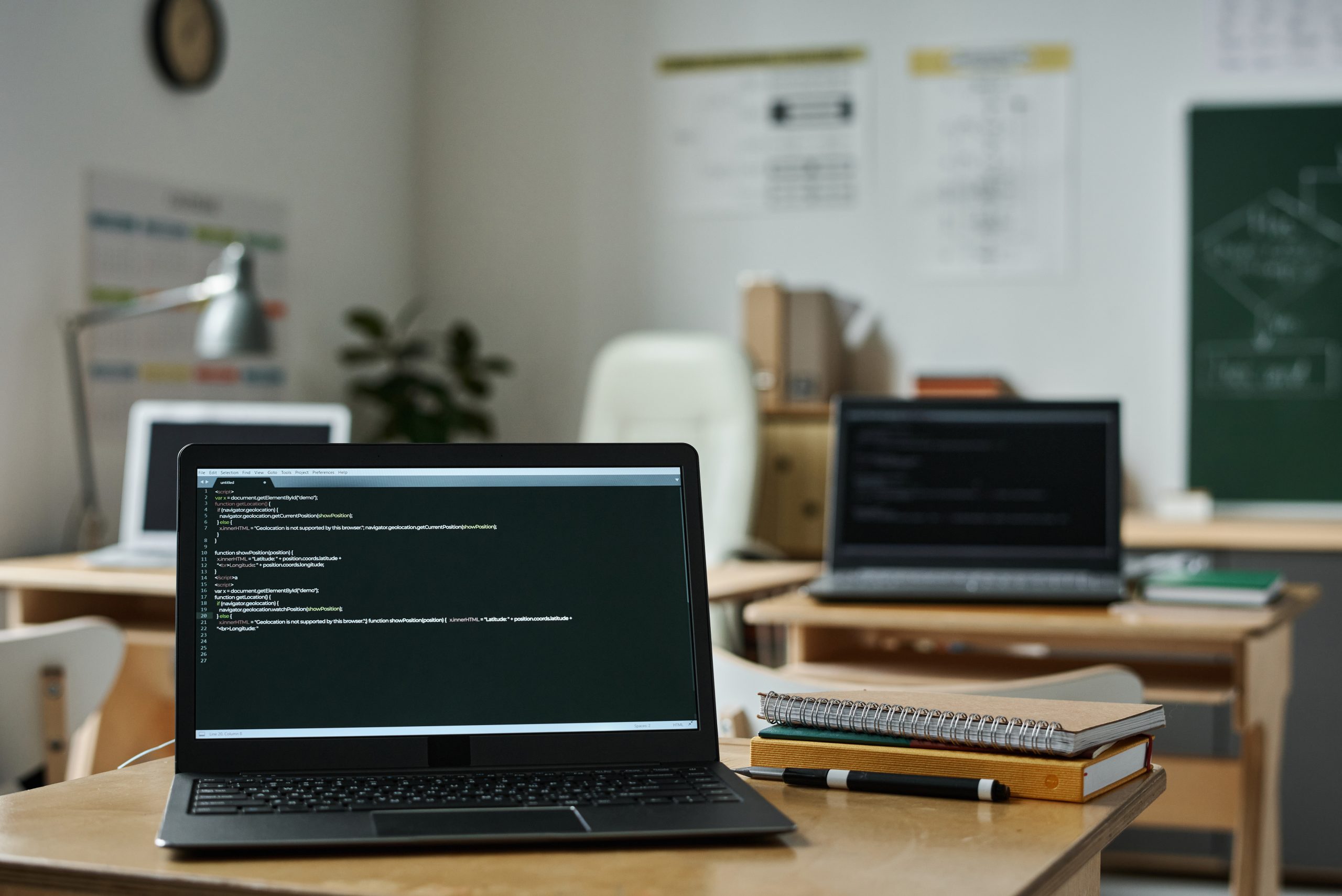 A laptop displaying code sits on a desk with notebooks and pens in a classroom. Other laptops and desks are visible, highlighting the need for electronics recycling for schools, alongside a chalkboard and posters on the walls.