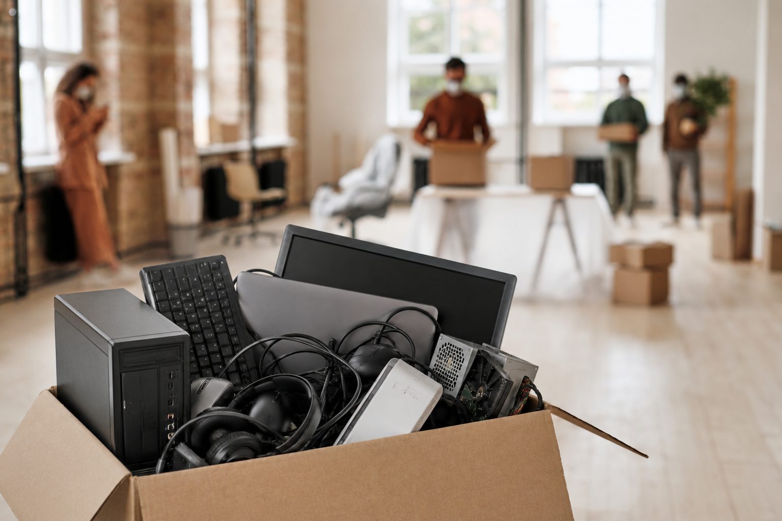 A cardboard box filled with computer equipment, such as a monitor, keyboard, cables, and headphones, is in the foreground of an office where people wearing masks are packing or carrying boxes for electronics recycling.
