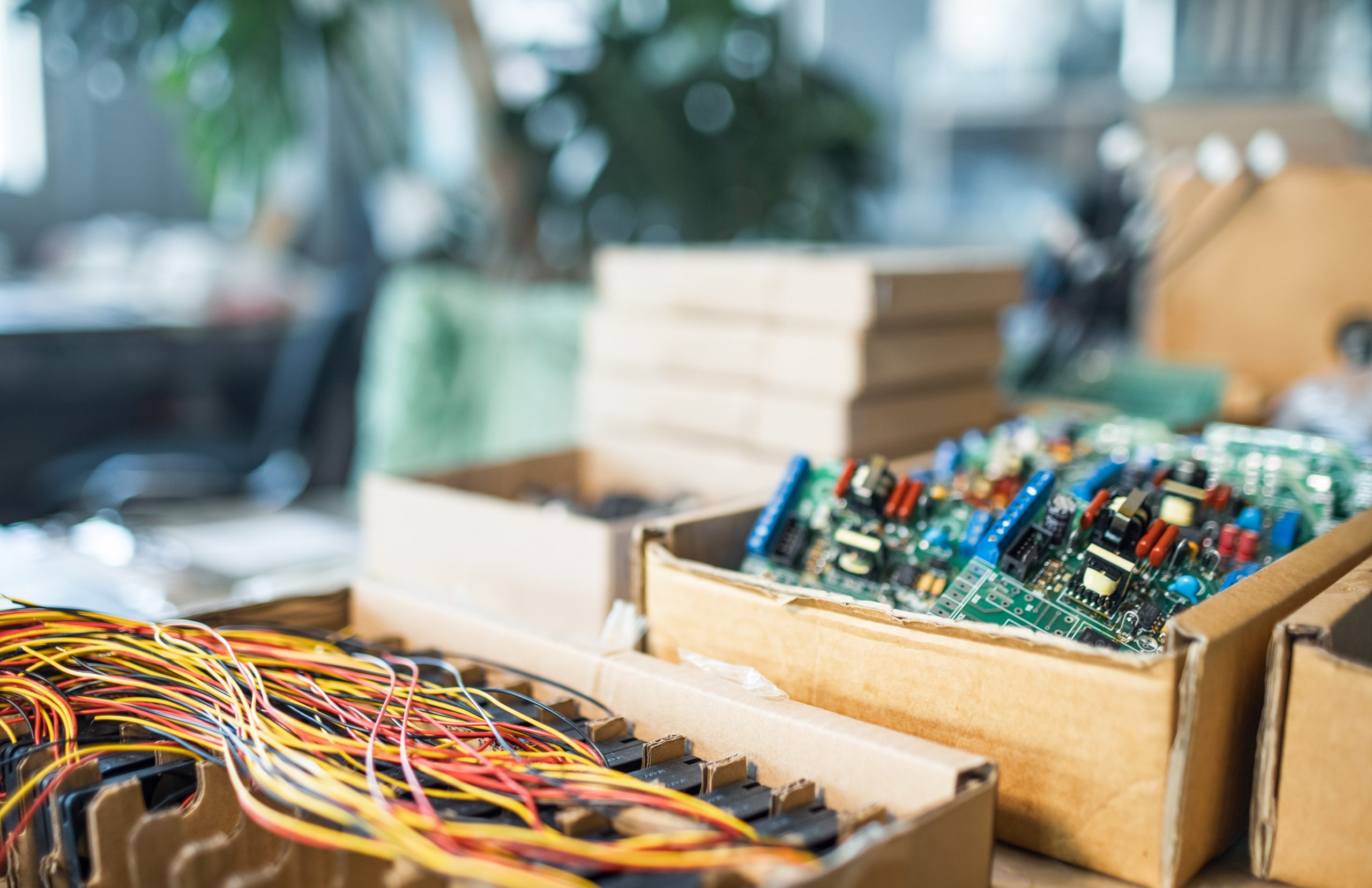 Close-up of cardboard boxes containing colorful electronic circuit boards and bundles of wires on a table, illustrating electronics recycling in a busy workspace or laboratory.
