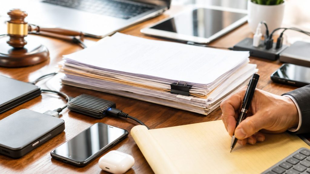 A person writes on a yellow legal pad at a cluttered desk with documents, a gavel, smartphone, hard drives awaiting electronic disposal, AirPods, a laptop, tablet, and a potted plant.