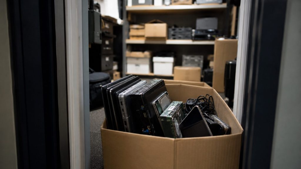 A cardboard box filled with old laptops and cables sits on the floor in a storage room with shelves holding various electronic equipment, cardboard boxes, and items awaiting data destruction.