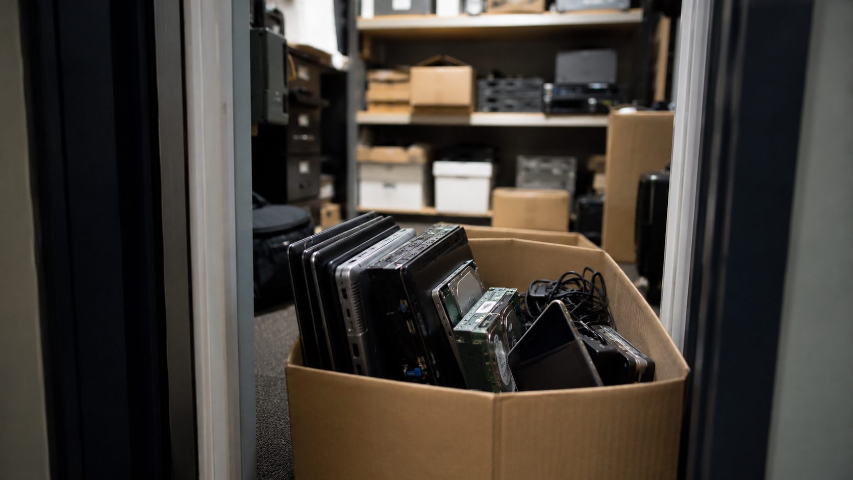 A cardboard box filled with old laptops and cables sits on the floor in a storage room with shelves holding various electronic equipment, cardboard boxes, and items awaiting data destruction.