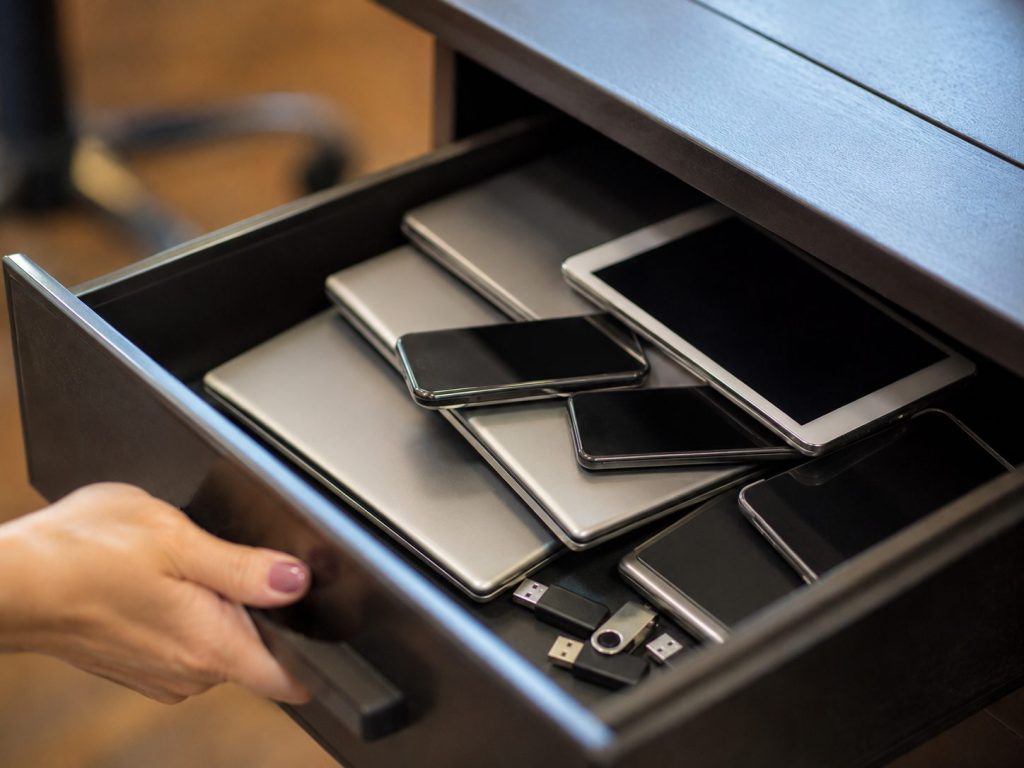 An open drawer filled with various electronic devices, including smartphones, tablets, laptops, and USB flash drives—prime candidates for data destruction. A hand is holding the drawer open.